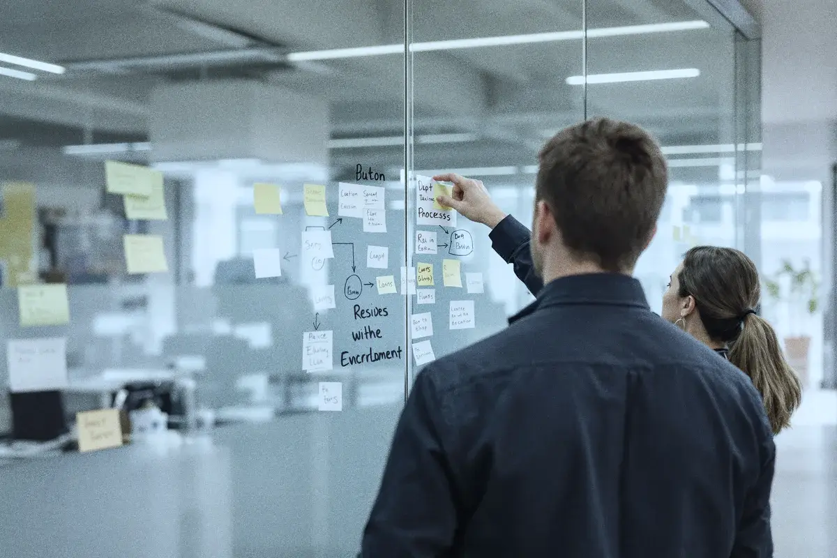 Two colleagues at a glass wall of sticky notes and a process flowchart